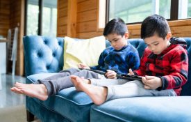 Two young boys sitting on a couch looking at tablets.