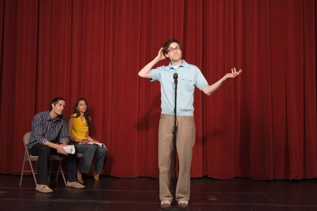 Performer in front of a microphone, doing poorly on his set while two people on stage watch from the side.