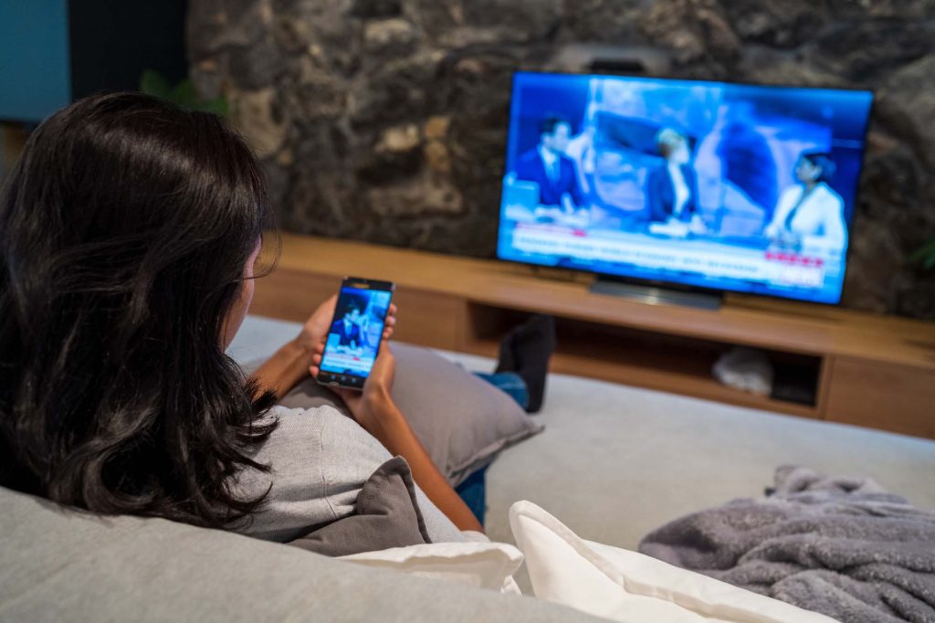 Woman uses her phone to watch news while news plays on the TV.