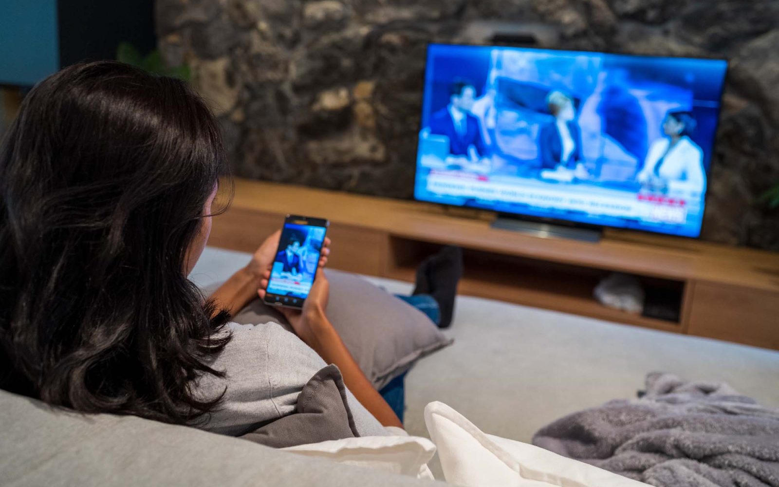 Woman uses her phone to watch news while news plays on the TV.